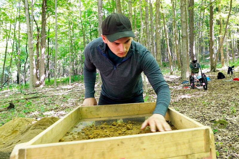 A college student works at an archaeology dig in the woods
